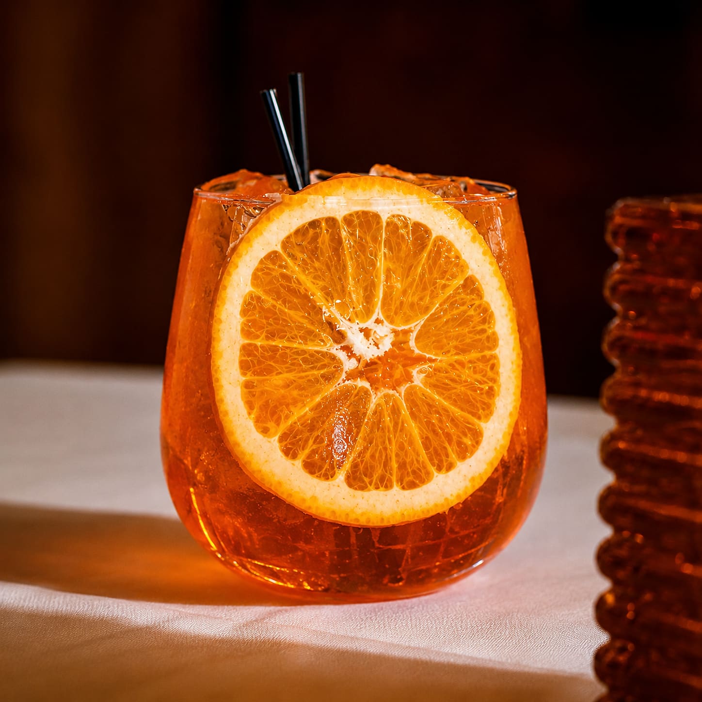 An Aperol spritz in a stemless glass with a wheel of orange in it, on a white tablecloth.