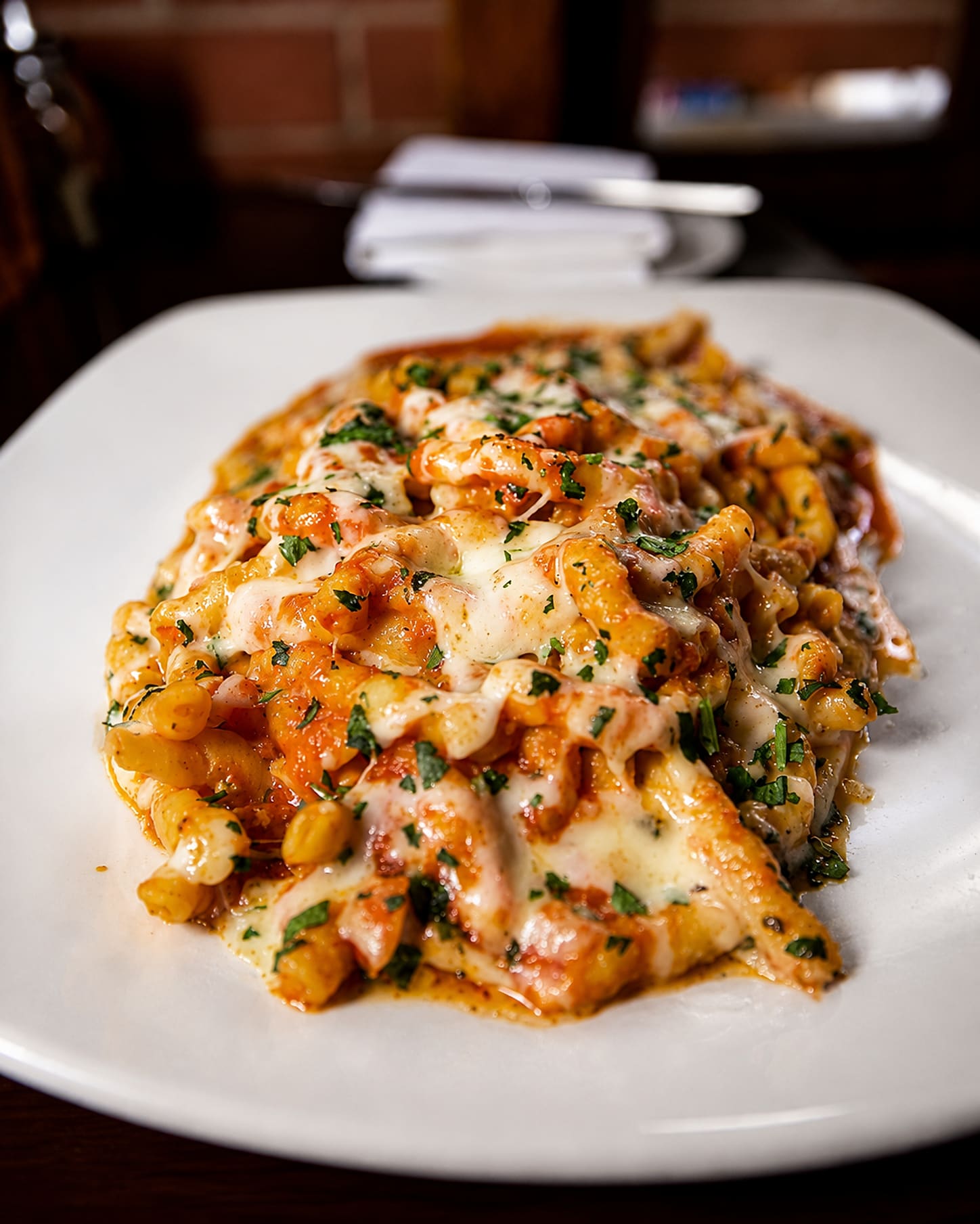 Baked pasta under melted mozzarella and tomato sauce on a white plate, with the restaurant’s brick wall behind it.