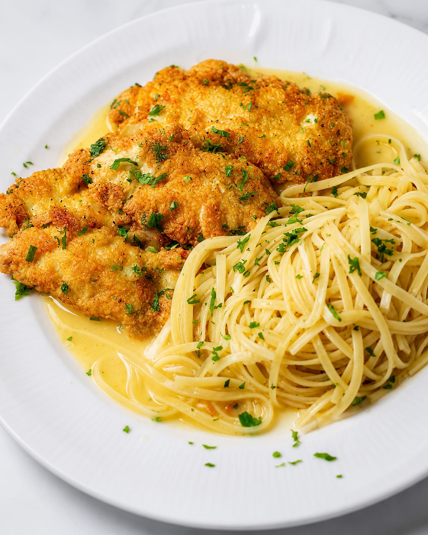 Golden egg-battered chicken cutlets in a lemon butter sauce beside a coil of linguini on a white plate, scattered with parsley.