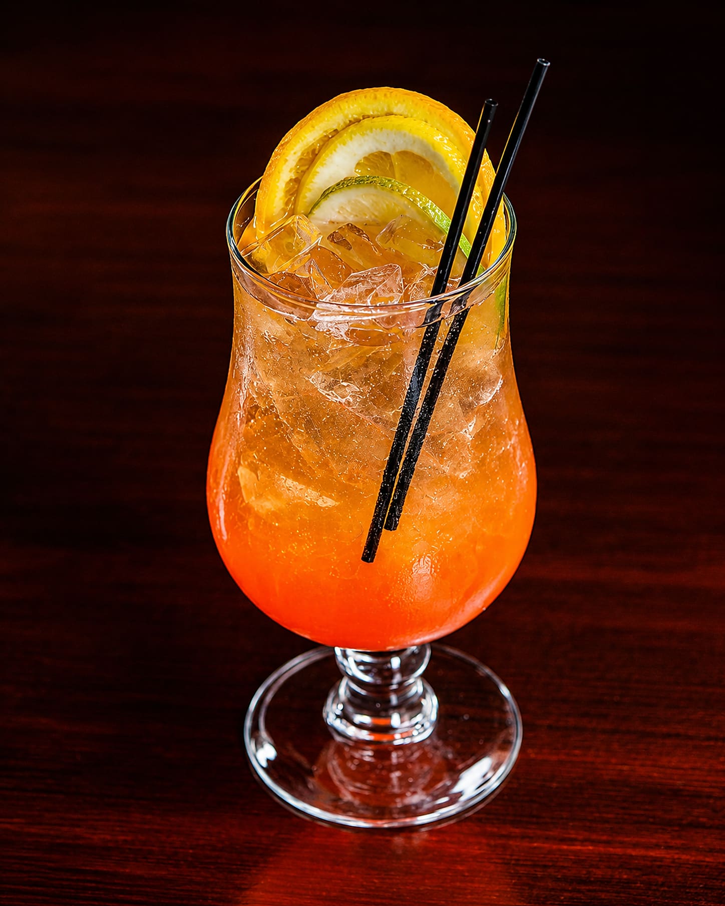 A tall citrus cocktail over crushed ice with orange, lemon and lime slices and two black straws, on a dark wood table.