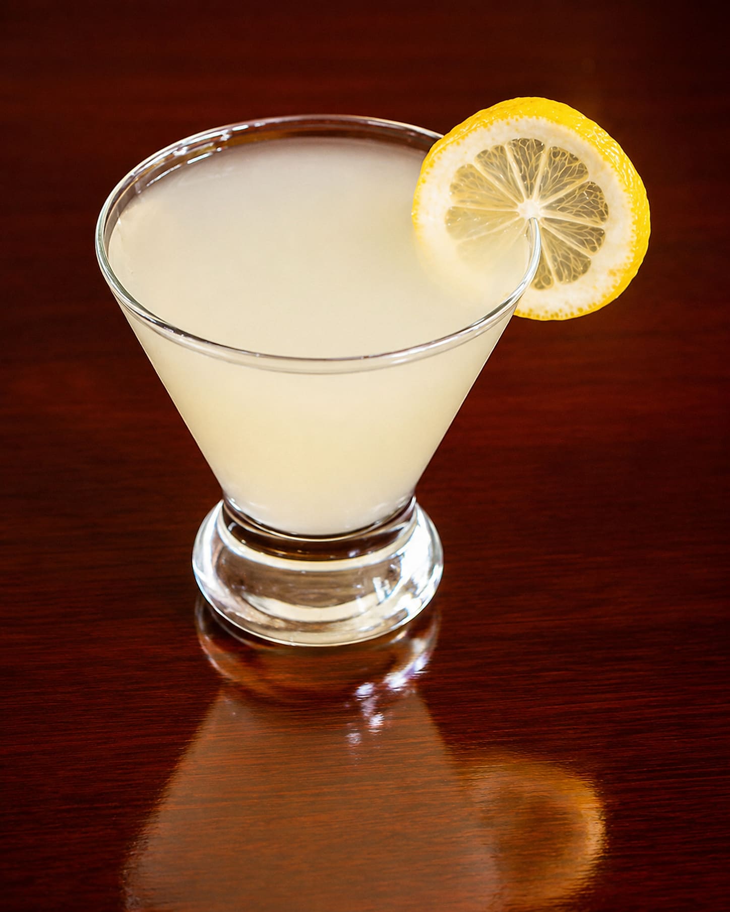 A lemon drop cocktail in a stemmed coupe with a wheel of lemon on the rim, reflected in a polished dark wood bar.