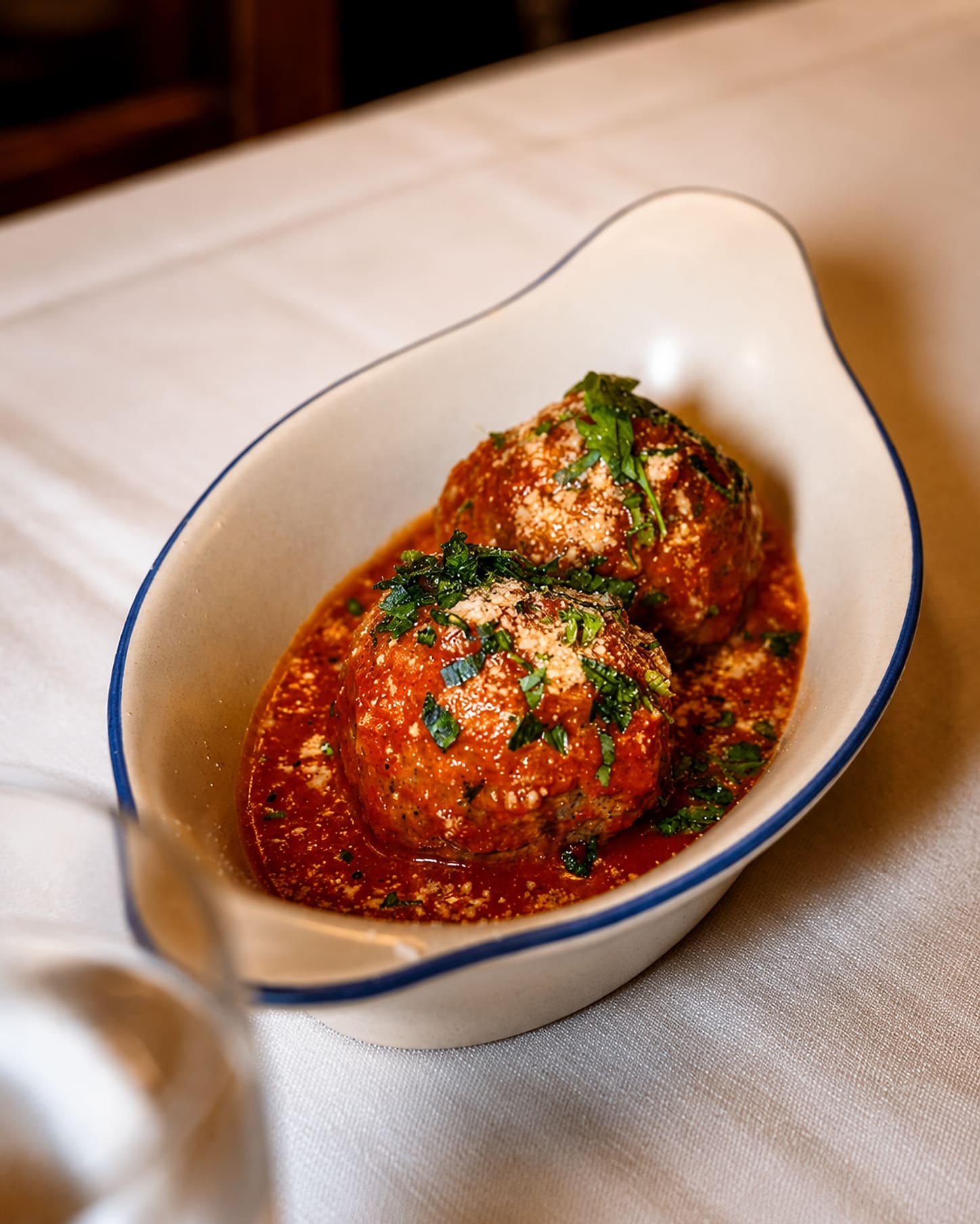 Two large meatballs in tomato sauce in a blue-rimmed oval dish, dusted with parmesan and chopped herbs, on a white tablecloth.