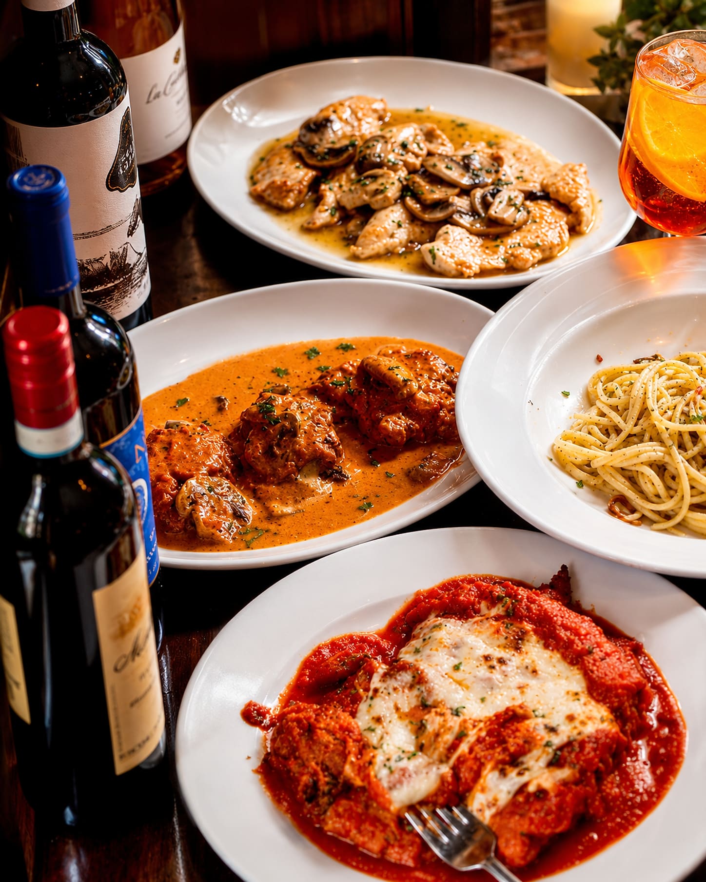 Four plates crowded onto one table — chicken with mushrooms in a white wine sauce, veal in a pink cream sauce, spaghetti with garlic and oil, and a chicken parmigiana — with open wine bottles and a spritz alongside.